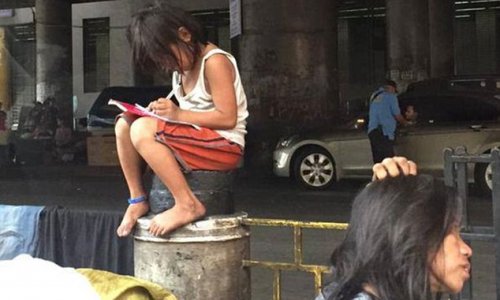 Girl hunches over her homework while sitting on a post alongside a filthy road